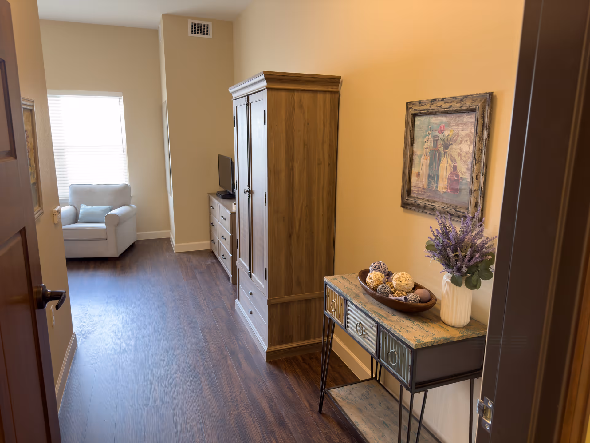 Entryway of a senior living suite with wood floors, a wooden armoire and dresser, a small armchair by the window, and a console table with a vase and decorative bowl.