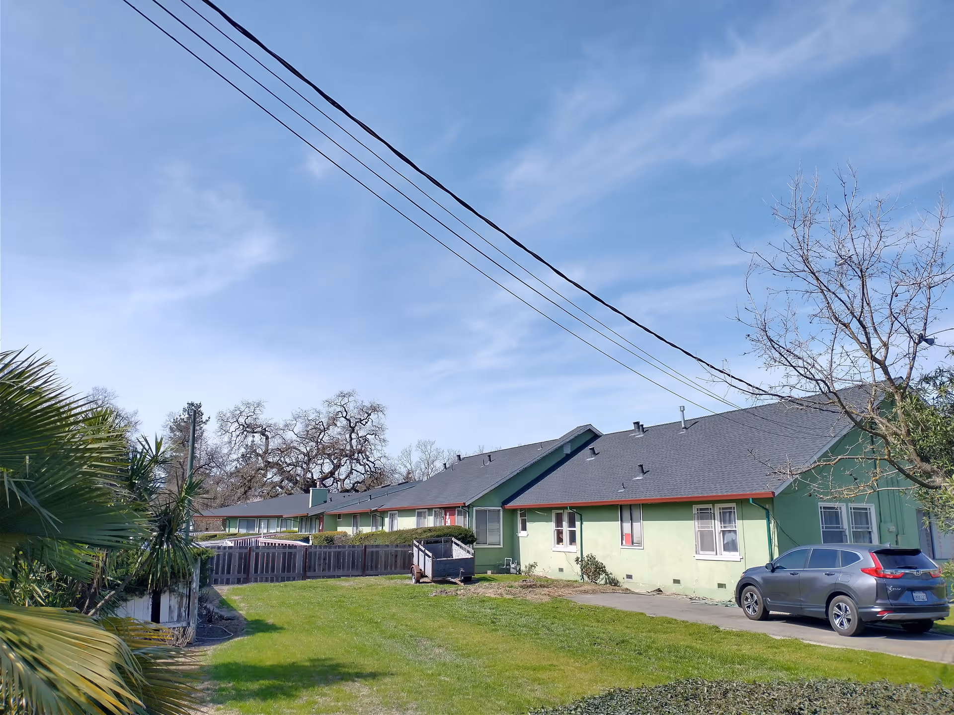 Exterior view of a single-story green building with a dark roof, a parked gray SUV, a small trailer, and a grassy area with trees and shrubs under a blue sky with some clouds.