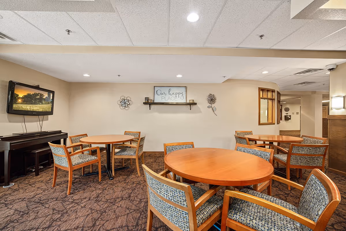 A cozy common area with three round wooden tables surrounded by patterned cushioned chairs. A piano is positioned against the left wall beneath a mounted flat-screen TV displaying a nature scene. The beige walls are decorated with a shelf holding candles and a sign that reads 'Our happy PLACE,' along with two metal floral wall art pieces. The carpet has a brown pattern, and the ceiling has recessed lighting.