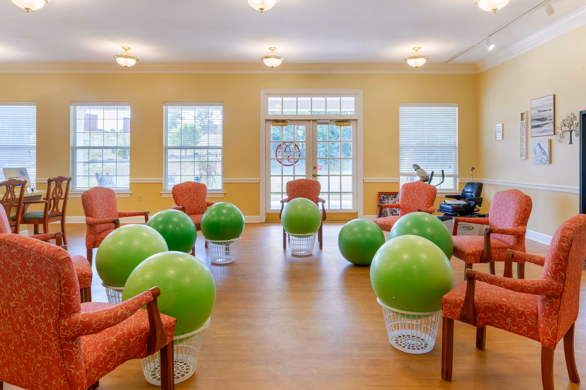 A bright activity room with red upholstered chairs arranged in a circle and large green exercise balls resting on white baskets, with windows and double glass doors at the far wall.