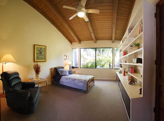 A cozy bedroom with a single bed covered in a light purple bedspread, a green recliner chair, a small side table with a lamp and vase, a large window showing greenery outside, and a built-in white bookshelf filled with books and decorative items. The room has a high wooden ceiling with exposed beams and a ceiling fan.