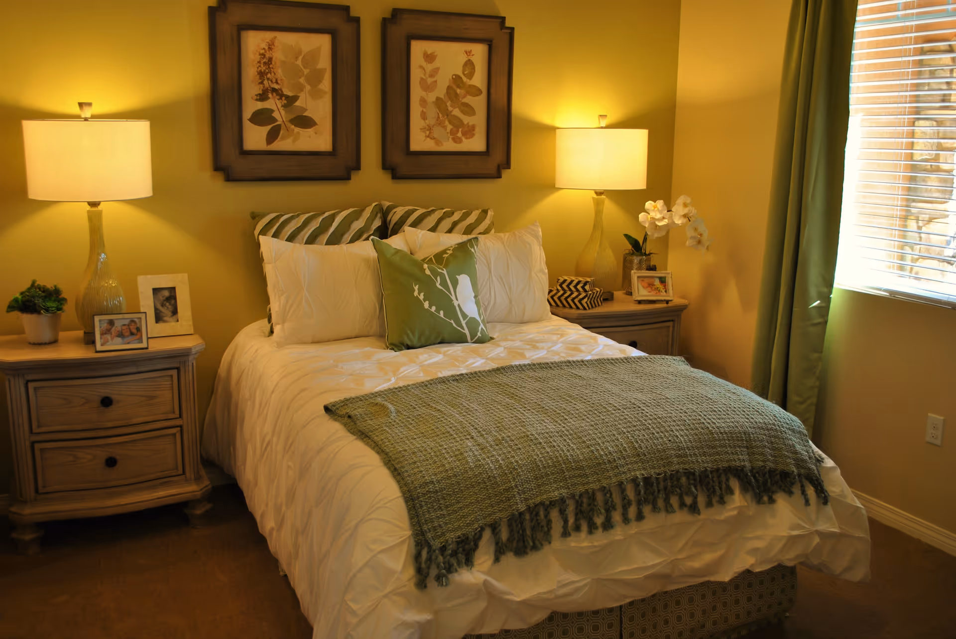 A cozy bedroom with a neatly made bed featuring white bedding, green and white pillows, and a green throw blanket. There are two wooden nightstands on either side of the bed, each with a lamp, framed photos, and decorative items. Two framed botanical prints hang on the wall above the bed. A window with green curtains allows natural light to enter the room.