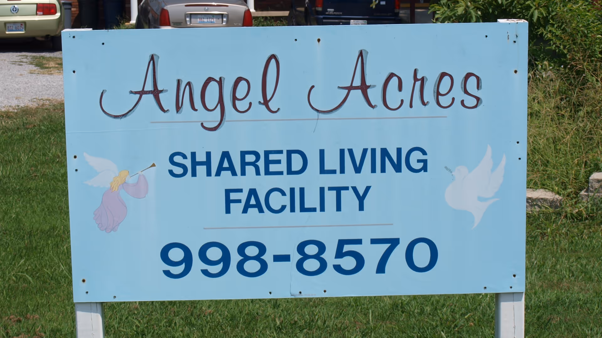 A light blue roadside sign reading 'Angel Acres SHARED LIVING FACILITY 998-8570' with small angel illustrations on a grassy lawn.