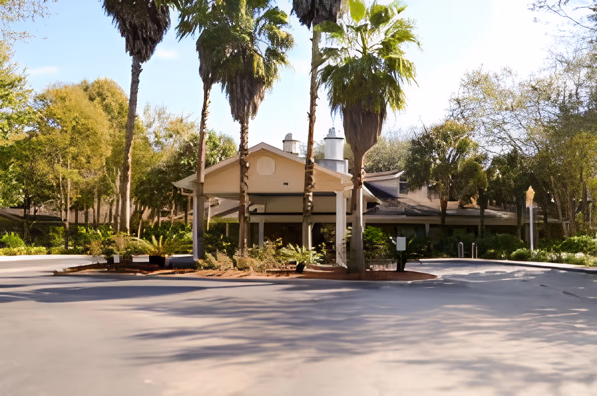 Front exterior view of Charter Senior Living of Gainesville building surrounded by tall palm trees and other greenery under a clear sky.