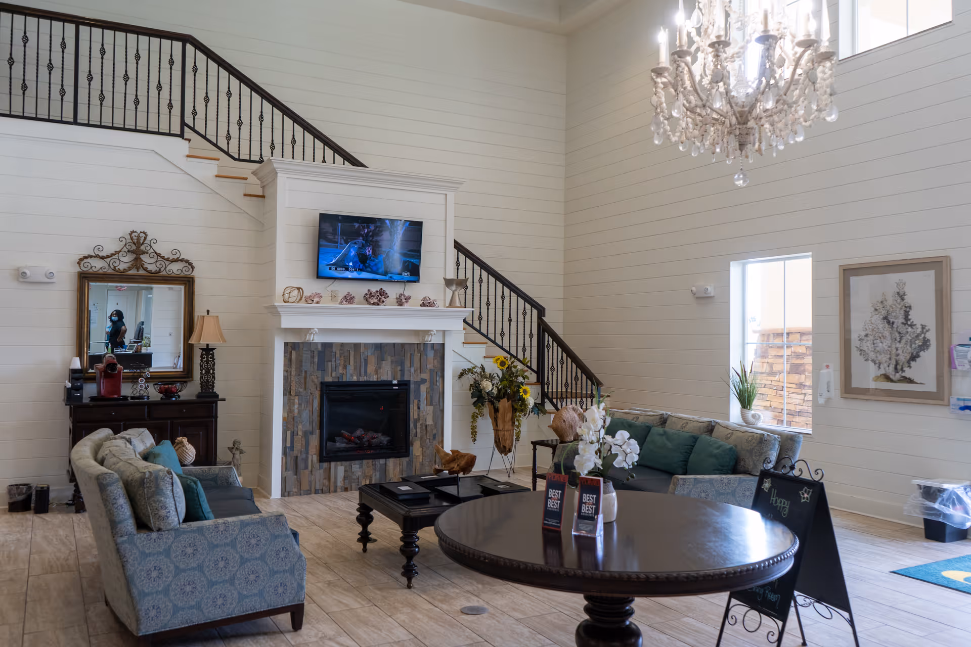 A spacious and well-lit living room with high ceilings, featuring a stone fireplace with a mounted TV above it, two patterned sofas with cushions, a round wooden table with flower arrangements, a chandelier, a staircase with black iron railing, a large mirror above a dark wooden cabinet, and wall art near a window.