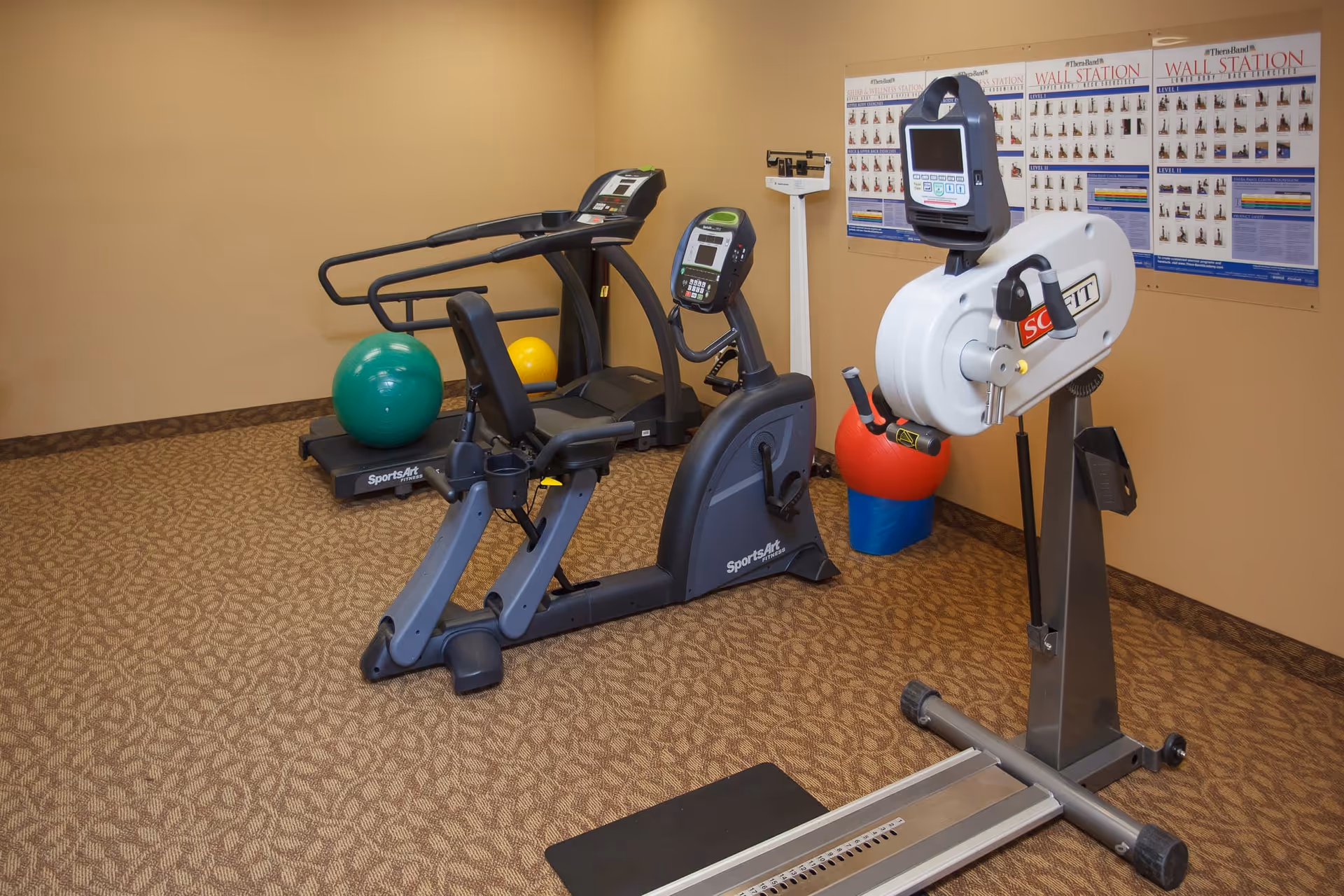 A small exercise room with fitness equipment including a treadmill, a recumbent exercise bike, a hand cycle machine, and colorful exercise balls. There are instructional posters on the beige walls and a scale in the corner.