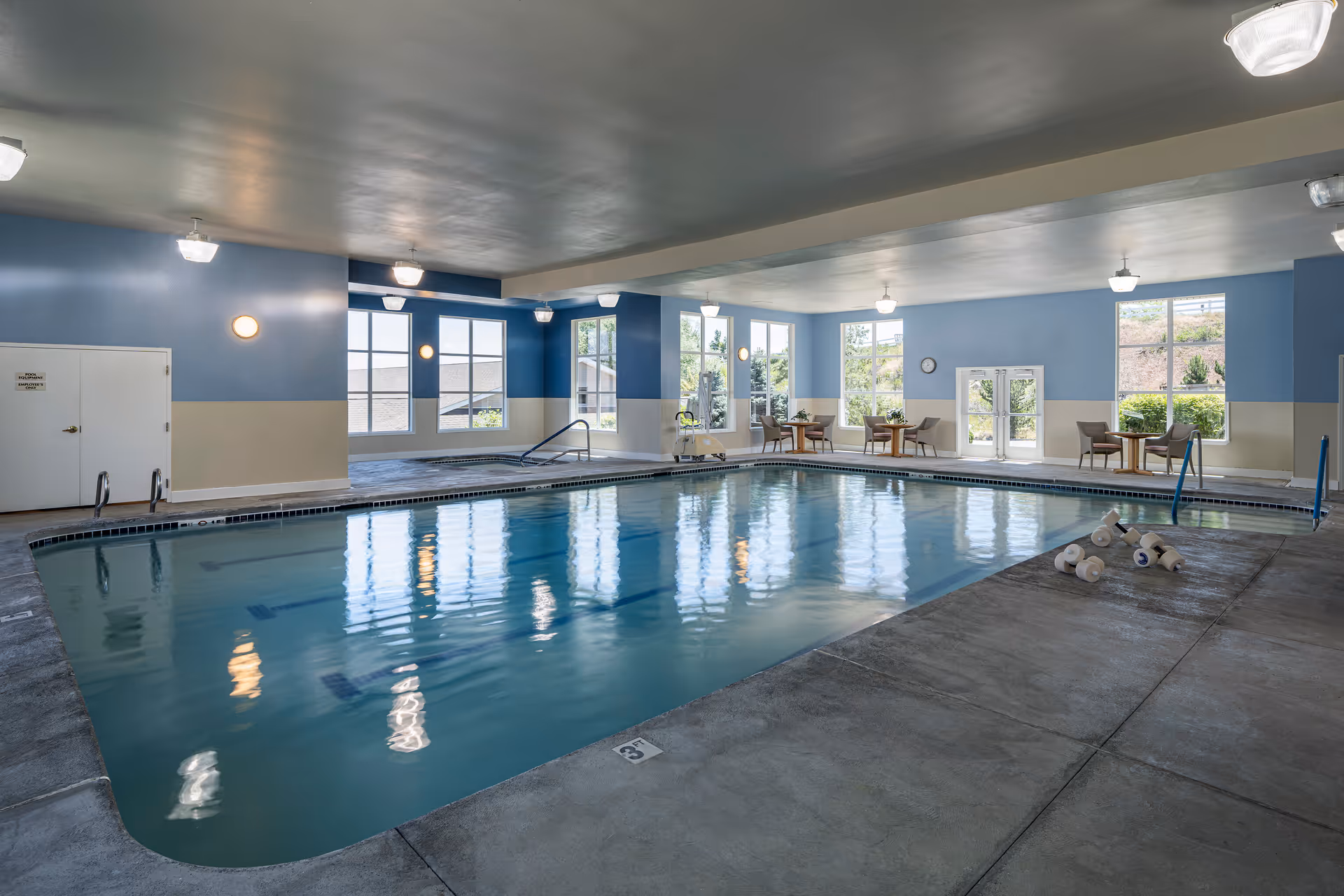 Indoor swimming pool area with blue and beige walls, large windows letting in natural light, several tables and chairs along the far wall, and pool equipment on the concrete floor near the pool edge.