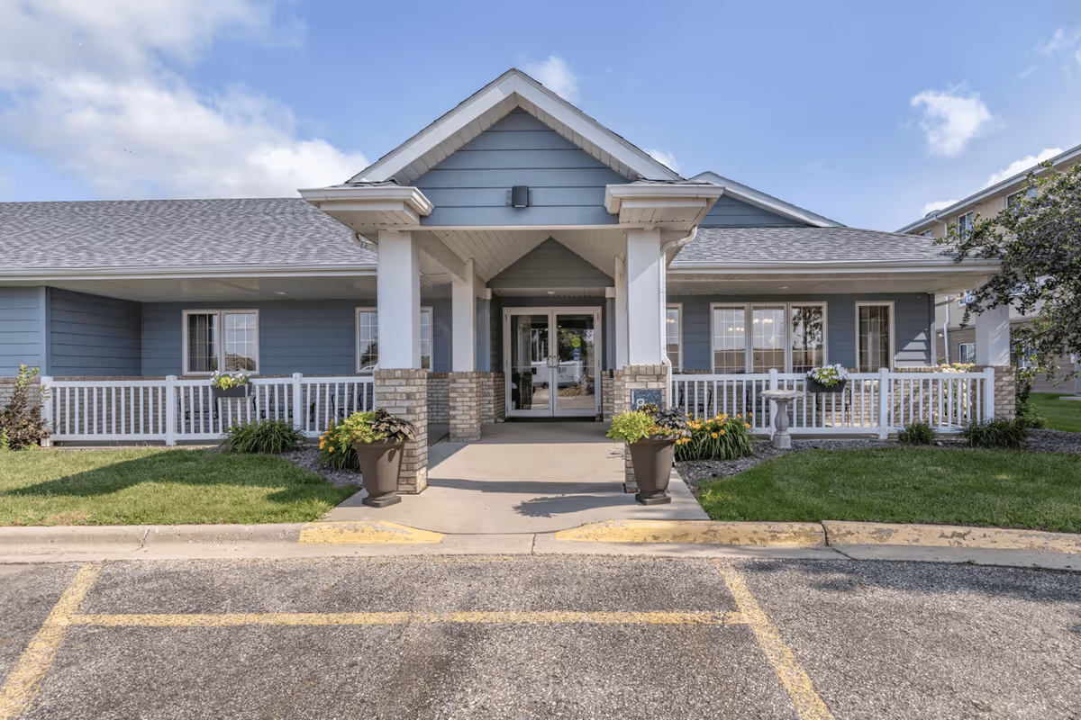 Front exterior view of Derian Place Senior Living building with a covered entrance, two large planters with flowers, white railing, and a well-maintained lawn under a partly cloudy sky.