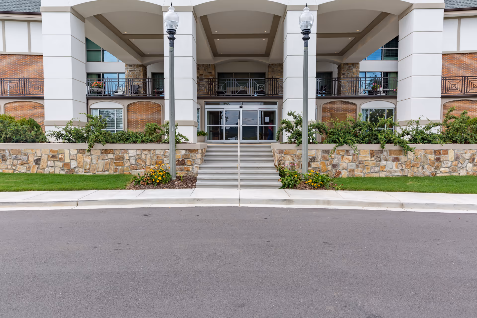 Front entrance of a senior living facility with stone and brick exterior, steps leading up to glass double doors, two lamp posts, and landscaped greenery with flowers.