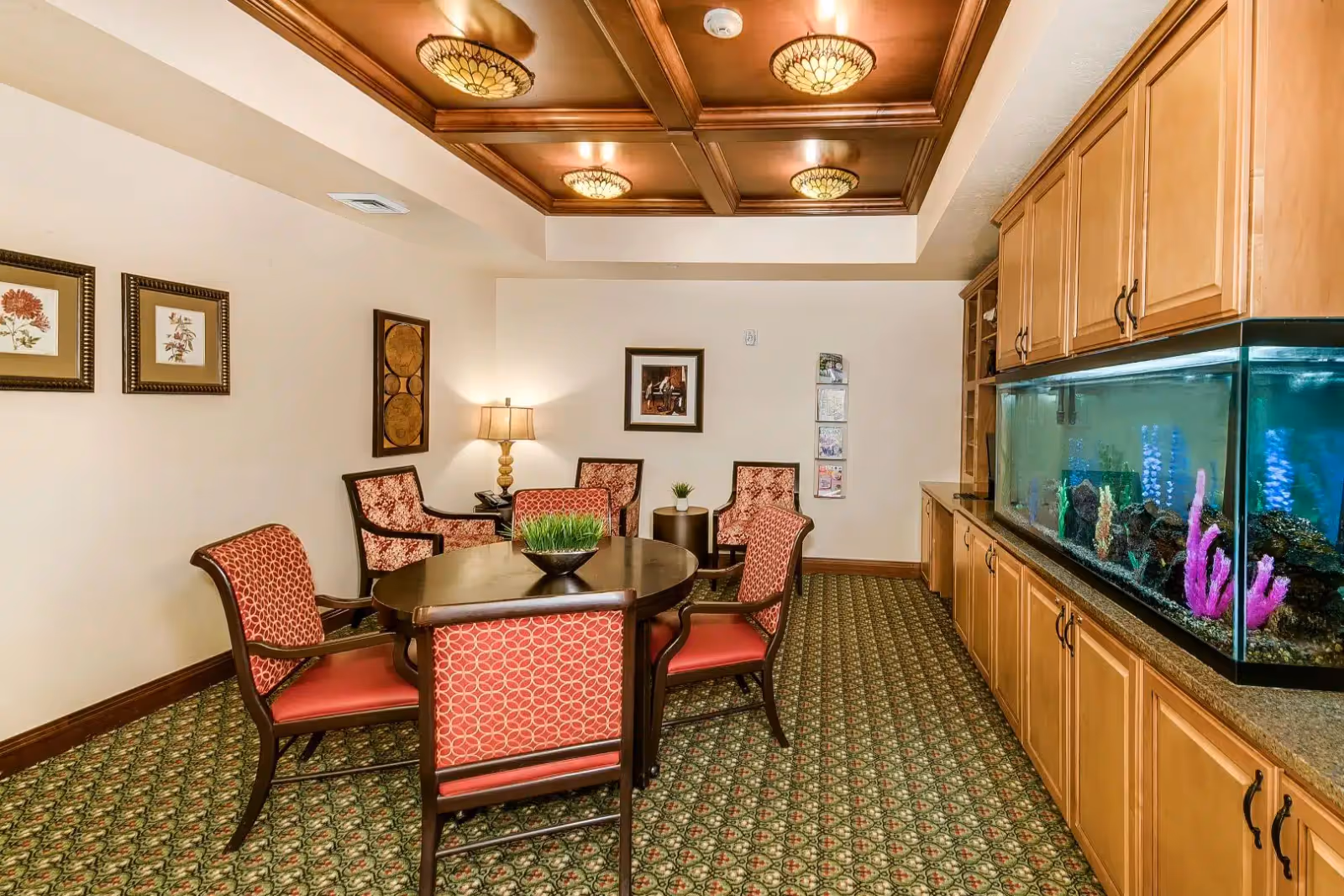 Cozy communal dining area with a round table surrounded by red upholstered chairs, decorative ceiling lights, framed artwork, and a large aquarium set into wooden cabinets.