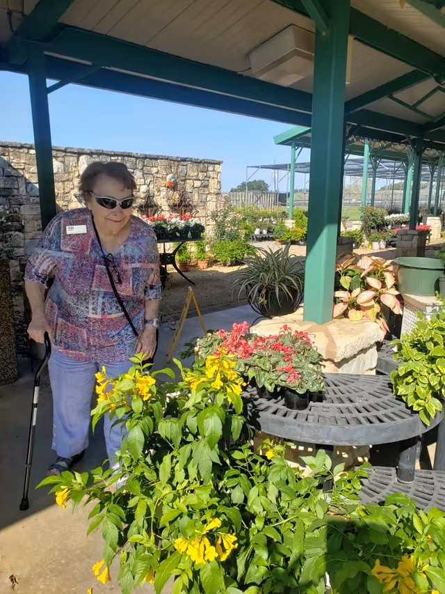 An elderly woman with a cane and sunglasses is standing under a covered outdoor area surrounded by various potted plants and flowers. The background shows a stone wall and more plants in a garden setting under a clear blue sky.