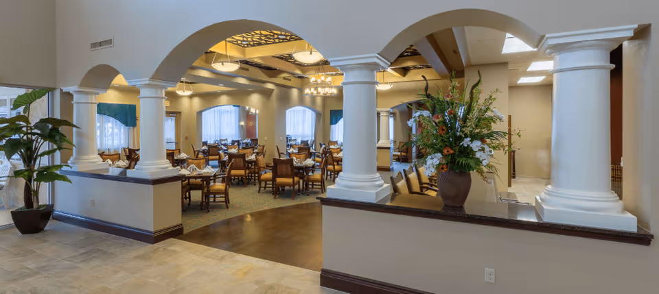 Spacious dining room with multiple tables and chairs arranged neatly, decorated with a large floral arrangement on a counter with white columns, warm lighting, and large windows with curtains.