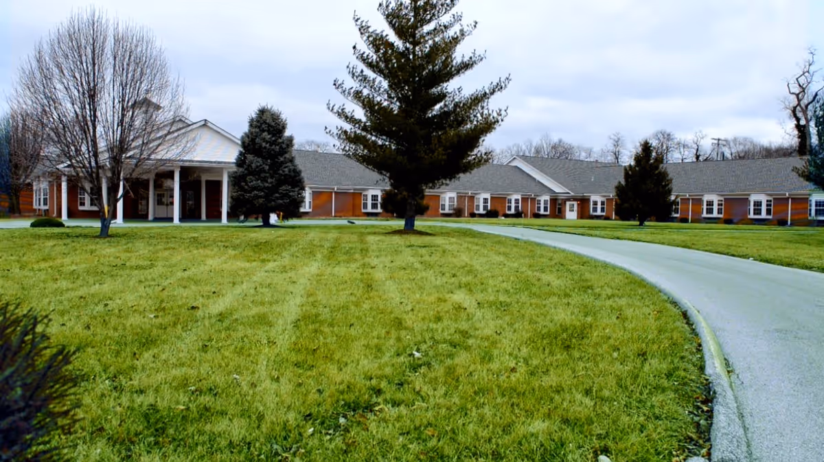 Wide view of Oakwood Estates Nursing & Rehab building with a large green lawn and a curved driveway leading to the entrance. The building is single-story with a gray roof and red brick walls, surrounded by several trees under a cloudy sky.