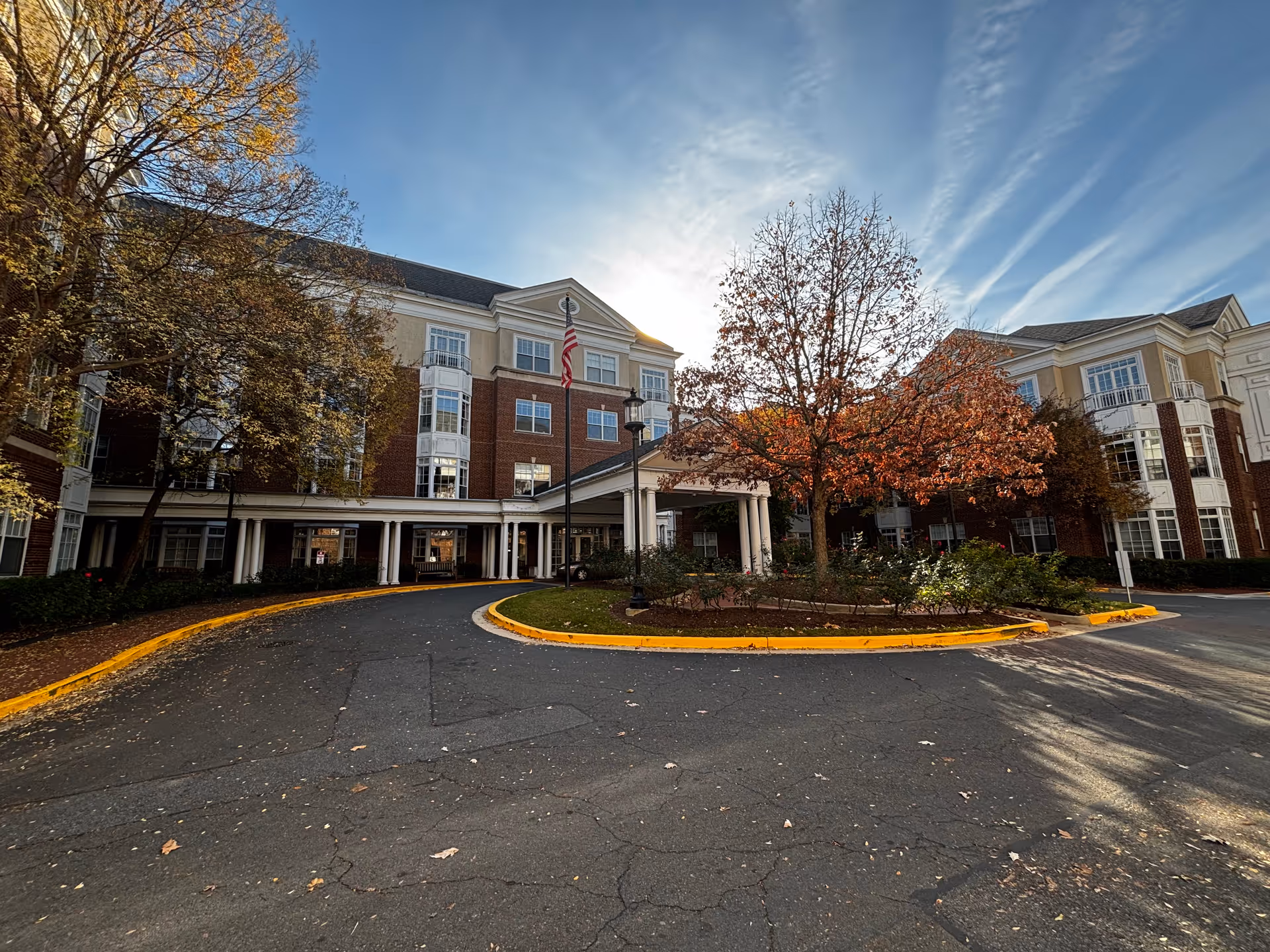 Front exterior view of a multi-story assisted living community building with a covered entrance, surrounded by trees with autumn foliage and a circular driveway.
