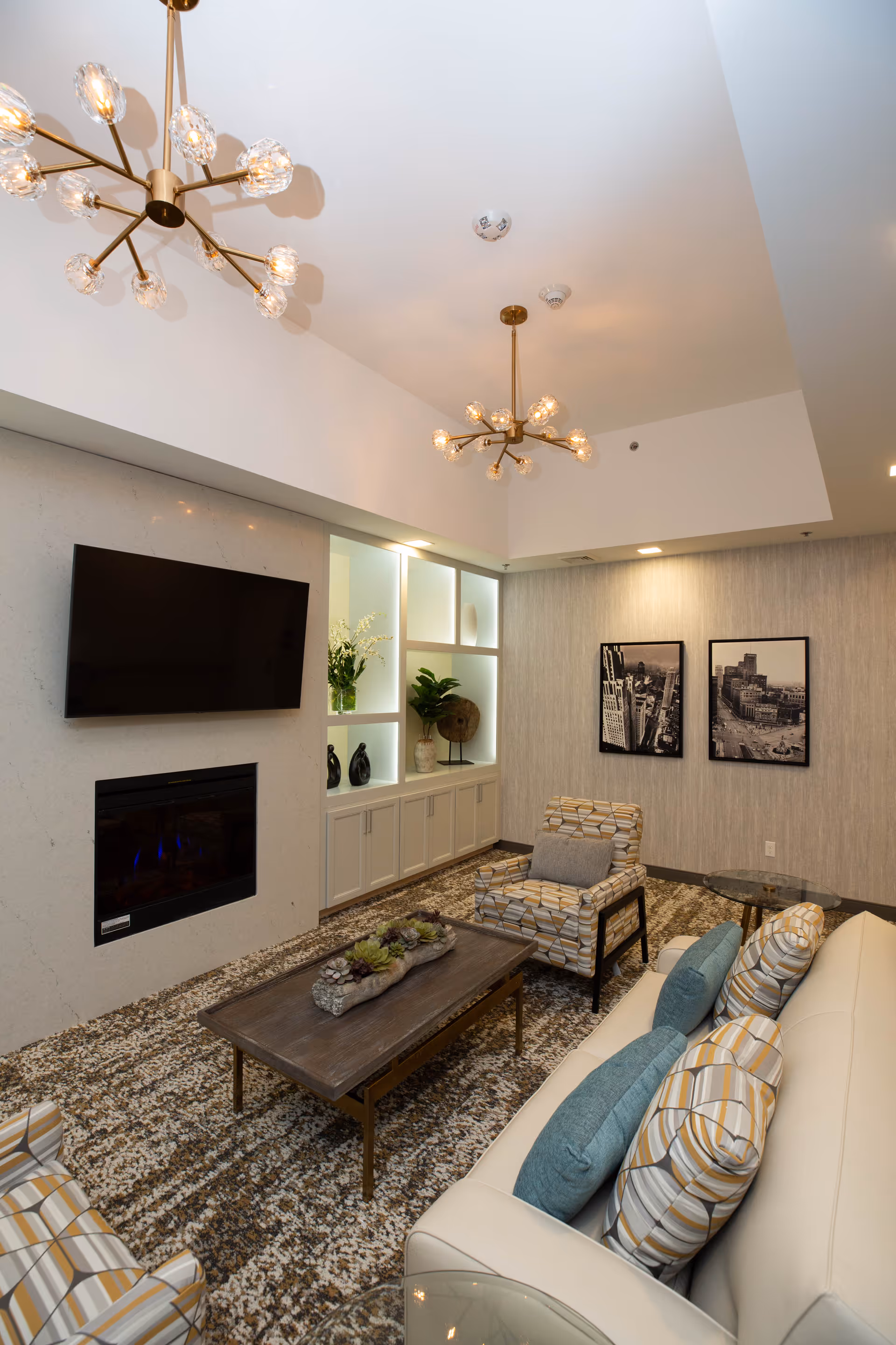 A modern living room with a white sofa adorned with patterned and solid blue pillows, two patterned armchairs, a wooden coffee table with a decorative centerpiece, a wall-mounted flat-screen TV above a built-in electric fireplace, and built-in shelving with plants and decorative items. The room features two contemporary chandeliers and two black-and-white framed cityscape photographs on the wall.