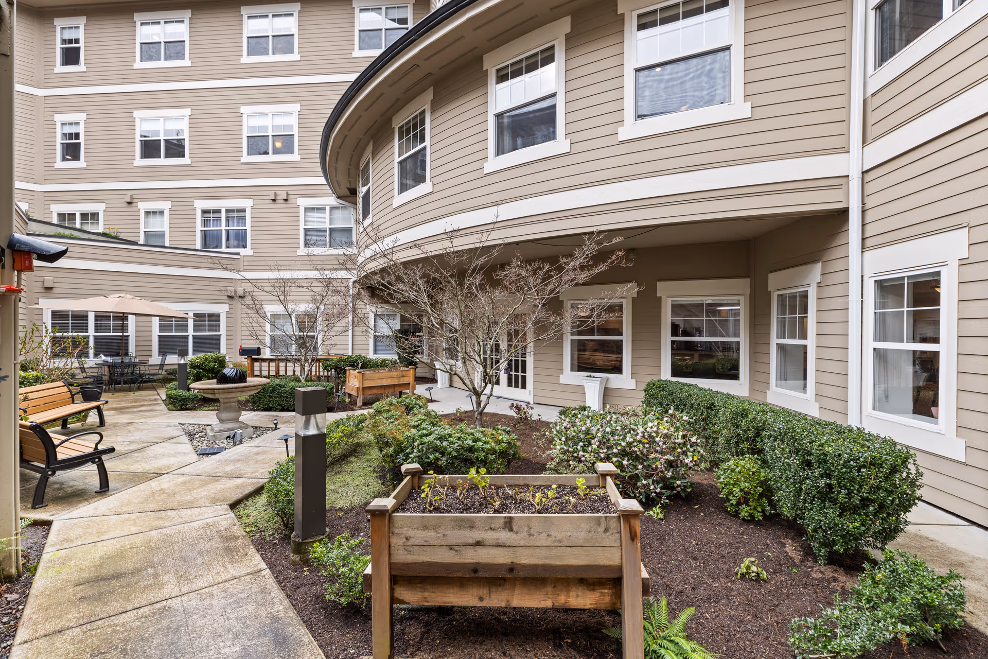 Outdoor courtyard area of a senior living facility with benches, a small fountain, planted shrubs, and a raised wooden planter box. The building surrounding the courtyard has multiple windows and beige siding with white trim.