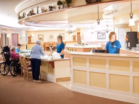 A bright and spacious reception area in a senior living facility with two staff members behind the counter assisting elderly residents, some seated and one in a wheelchair. The area features warm lighting, decorative items on a shelf above, and a welcoming atmosphere.
