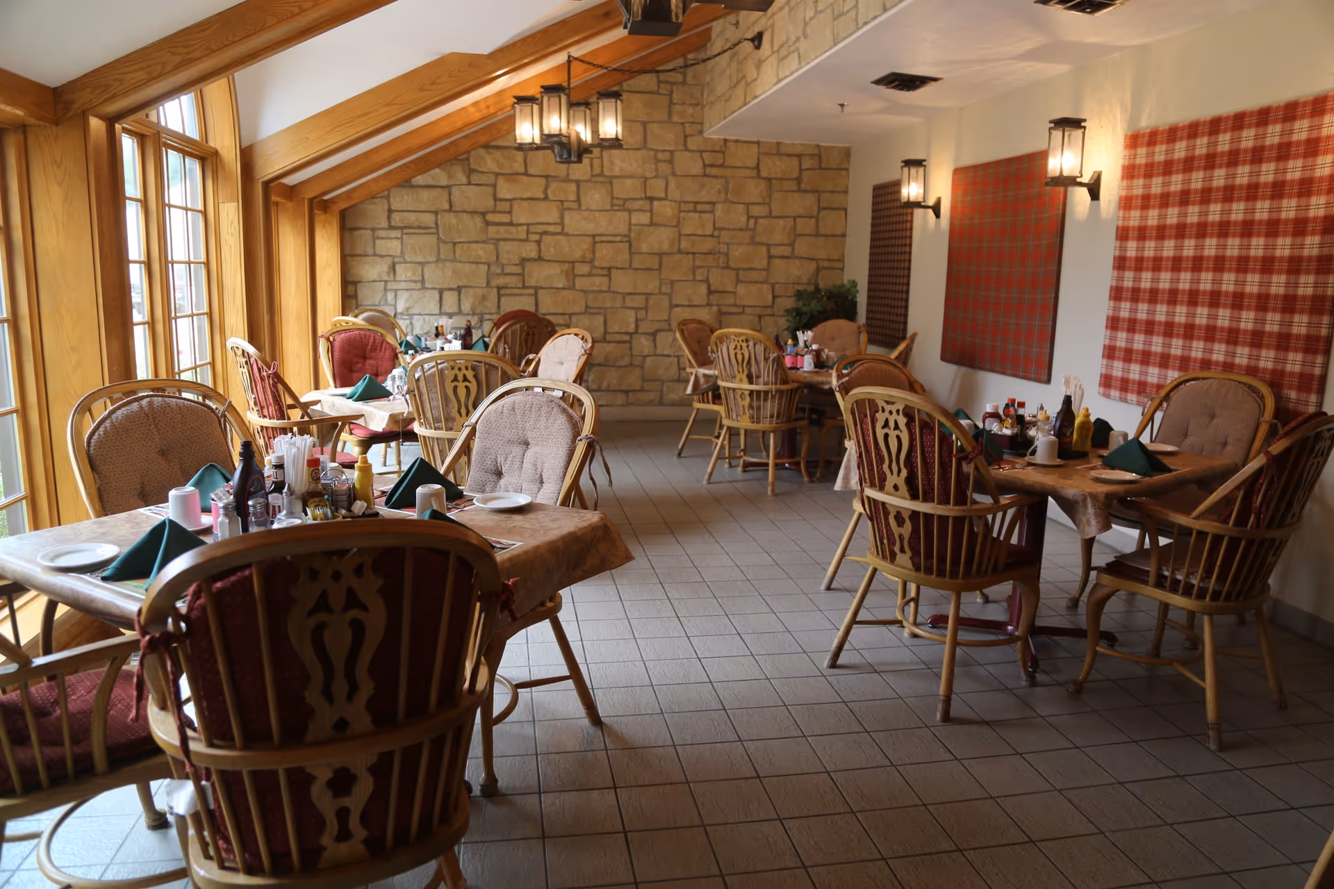 A cozy dining room with multiple wooden tables and cushioned chairs arranged neatly. The tables are set with plates, napkins, condiments, and utensils. The room features large windows on the left side allowing natural light to fill the space, a stone accent wall at the back, and plaid fabric wall decorations on the right. Warm lighting fixtures hang from the ceiling and are mounted on the walls.