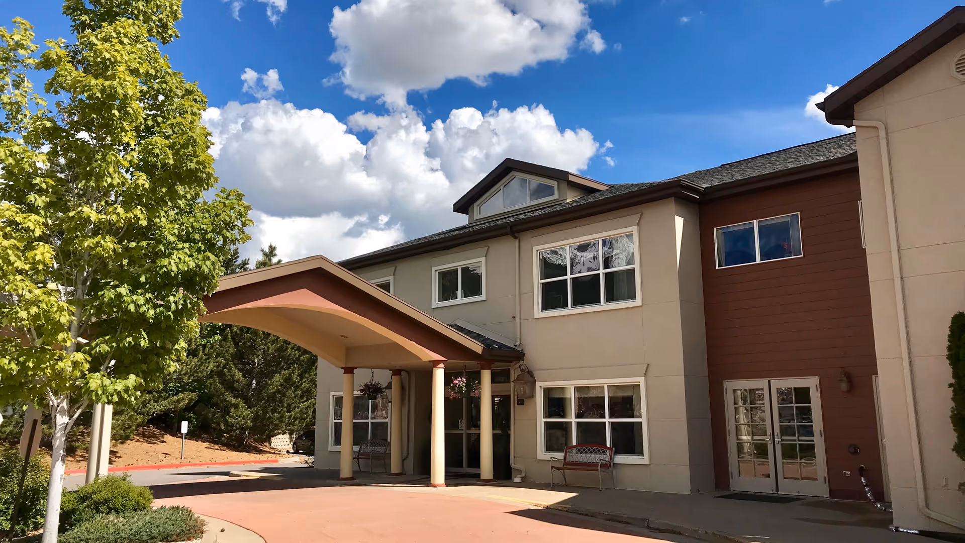Front entrance of a two-story building with a covered porte-cochere, benches, landscaping, and a blue sky with clouds.