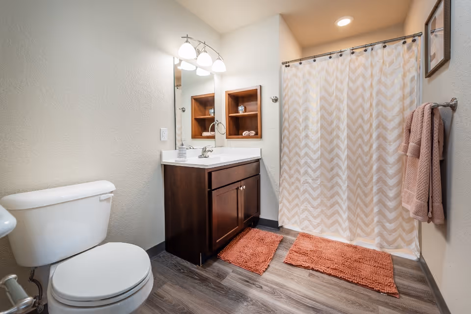 A clean and modern bathroom featuring a white toilet, a dark wood vanity with a white countertop and sink, a large mirror with three overhead lights, a built-in wooden shelf with decorative items, a shower with a chevron-patterned curtain, two orange bath mats on the floor, and a towel rack with folded towels on the wall.