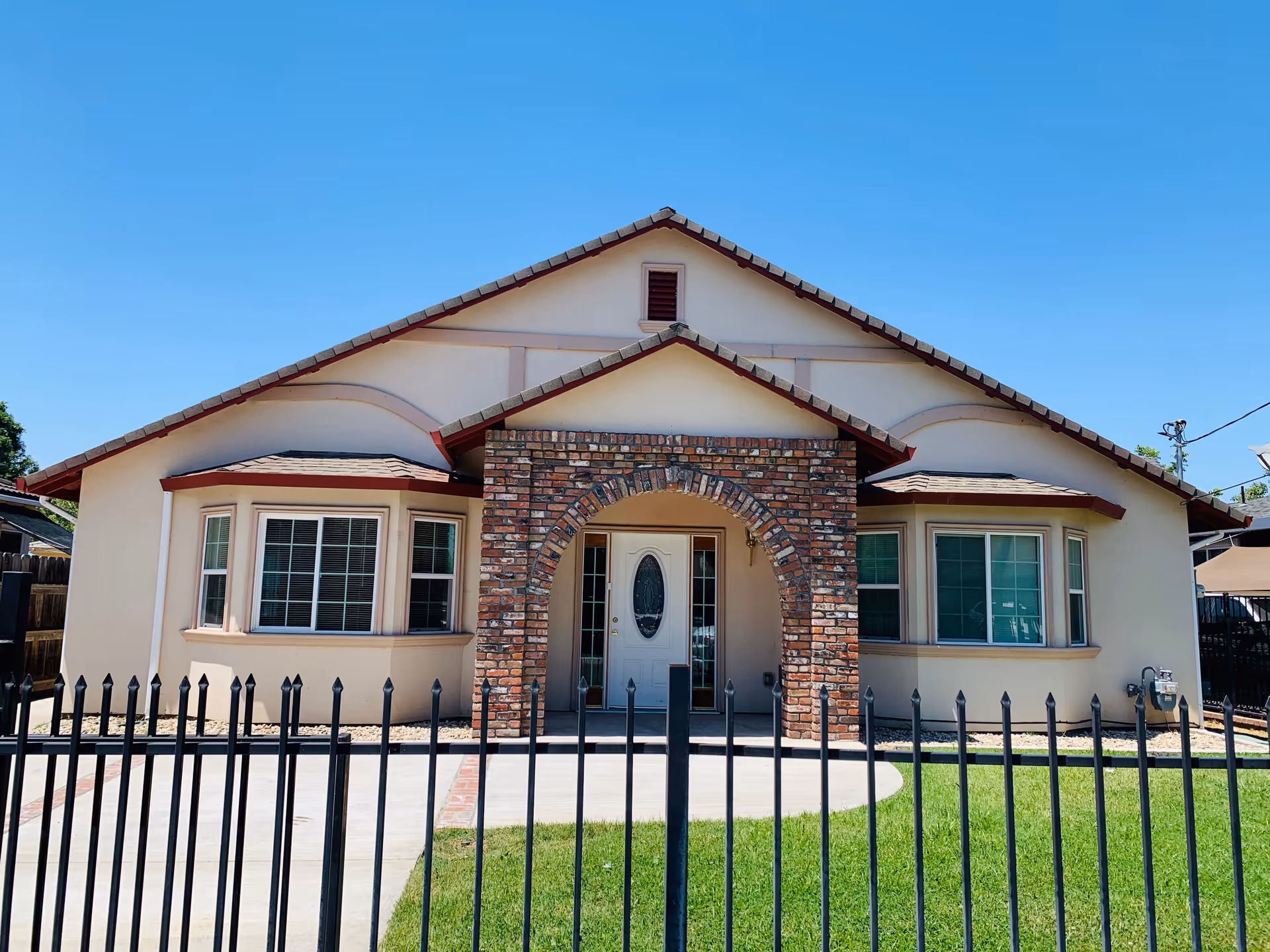 Front exterior view of a single-story assisted living facility building with a beige facade, two bay windows, a white front door with an oval glass panel, and a brick archway entrance. The building is surrounded by a black metal fence and a well-maintained green lawn under a clear blue sky.