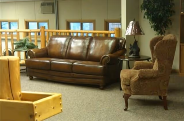 A cozy seating area with a brown leather sofa and a patterned upholstered armchair next to a small round table with a lamp. The background shows wooden railings and windows letting in natural light.