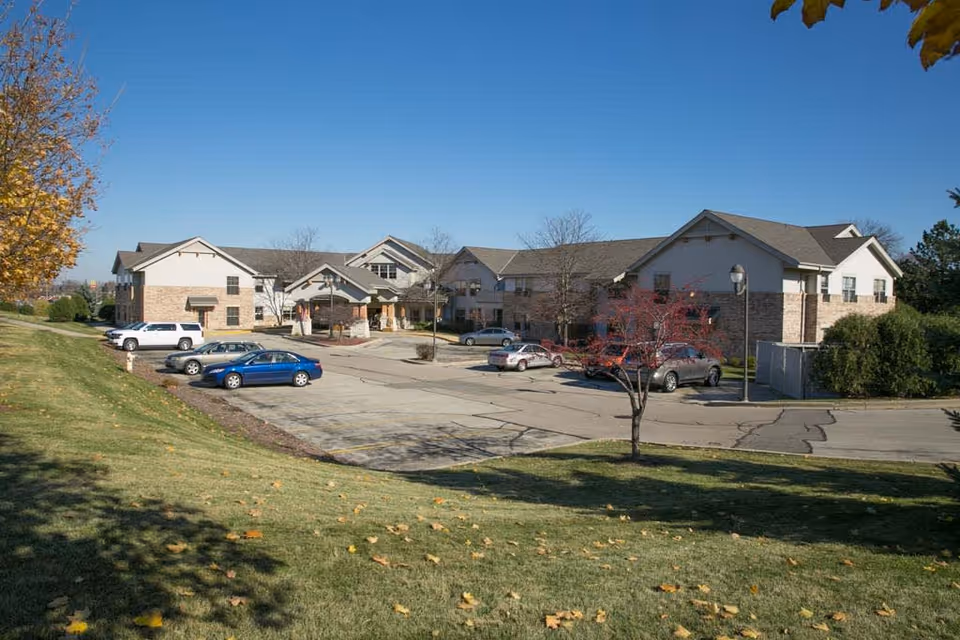Exterior view of Riverview Village Senior Living facility showing a two-story building with a parking lot in front, several parked cars, a small tree with red leaves, and a grassy area with scattered fallen leaves under a clear blue sky.