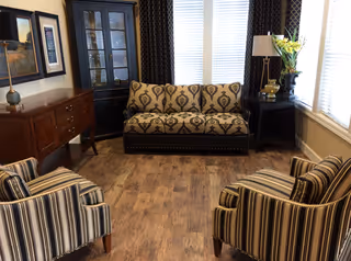 A cozy living room with a patterned sofa against a window with white blinds and dark curtains. Two striped armchairs face the sofa on a wooden floor. There is a wooden sideboard with framed artwork above it on the left, a black corner cabinet, and a side table with a lamp and a vase of yellow flowers on the right.