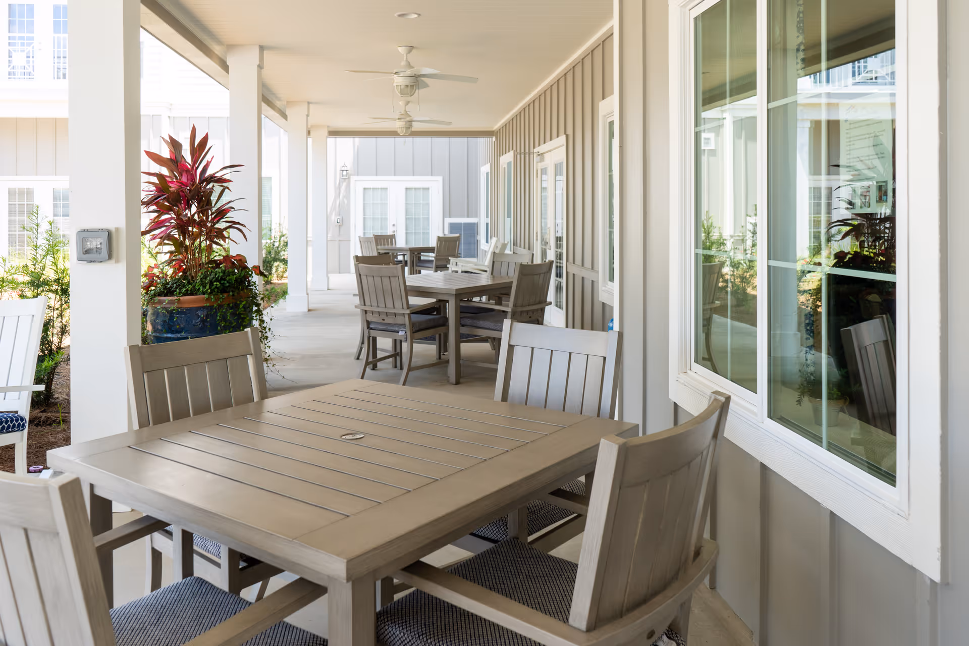 Covered outdoor patio area with multiple wooden tables and chairs arranged for seating. The patio is adjacent to a building with large windows and light-colored siding. There are ceiling fans mounted on the patio ceiling and potted plants adding greenery to the space.
