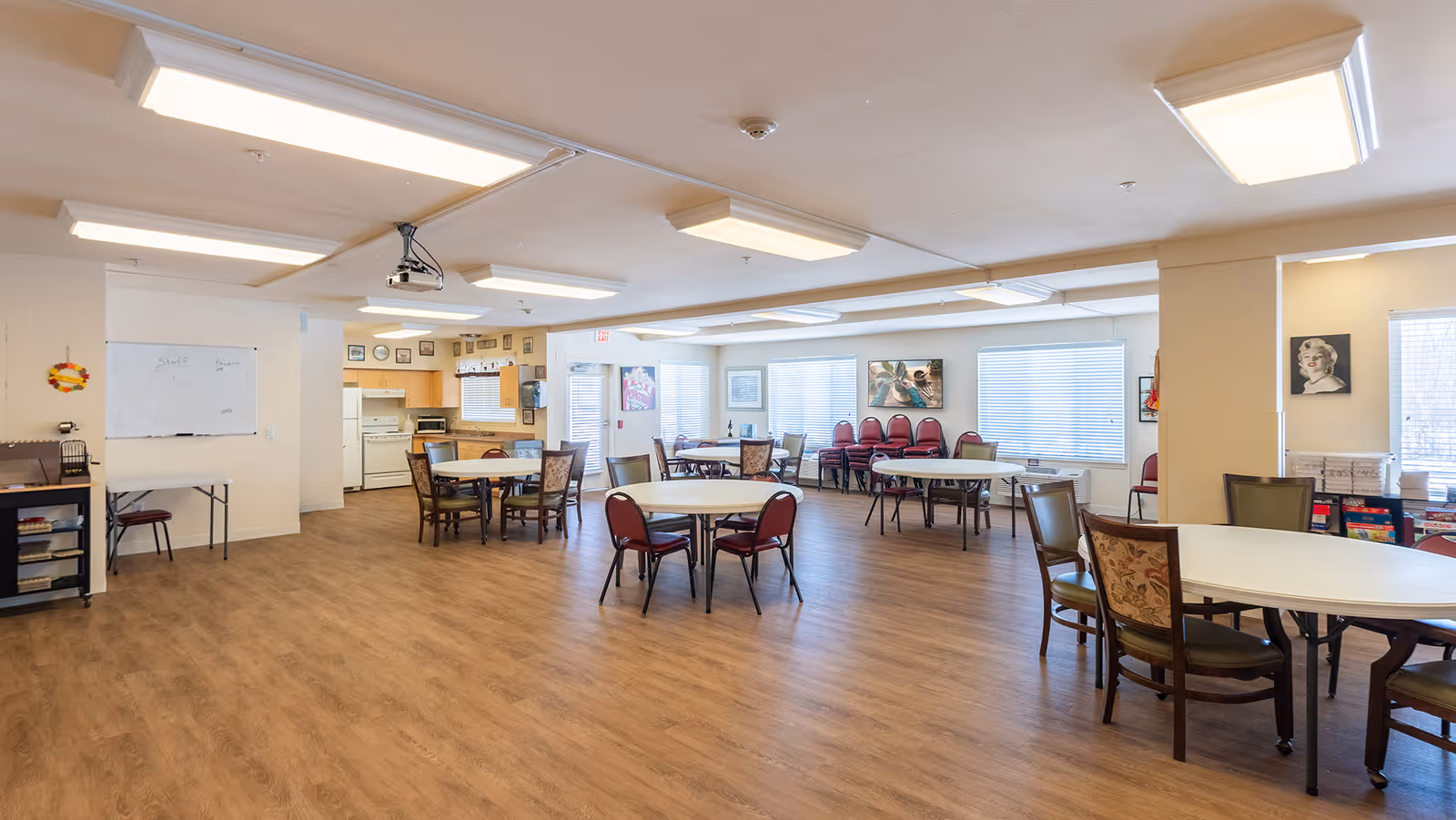 A spacious common area in a senior living facility with multiple round tables and chairs arranged throughout the room. The room has wood flooring, large windows with blinds, and bright overhead lighting. A small kitchen area with appliances is visible in the background, along with some stacked chairs and framed artwork on the walls.