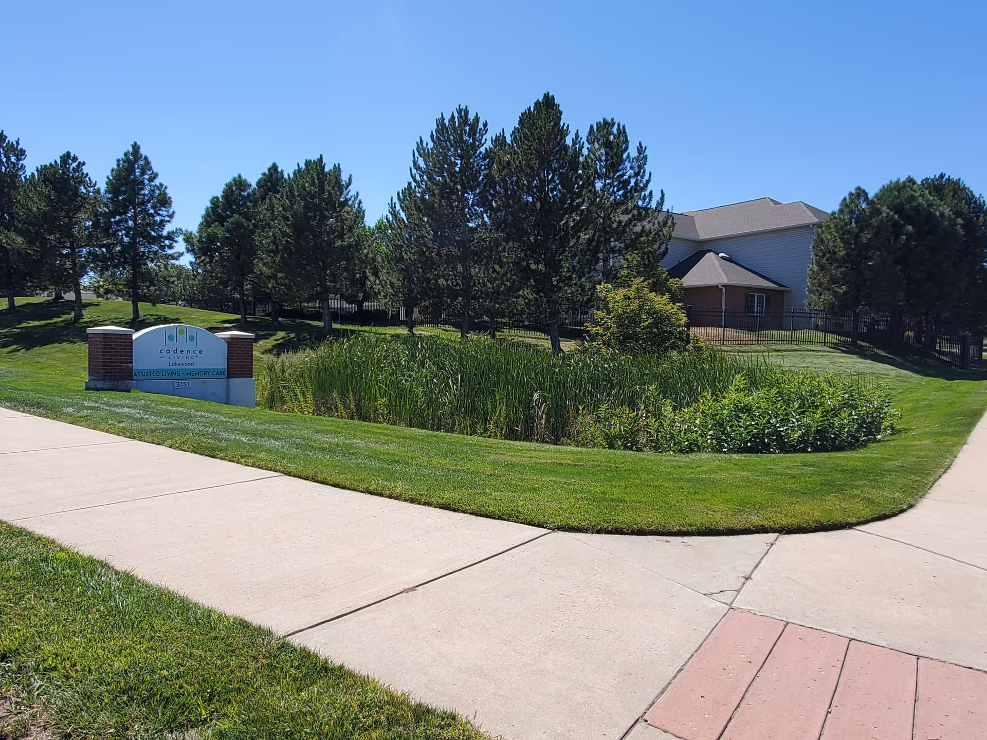 Outdoor view of a senior living facility with a well-maintained lawn, a small pond surrounded by tall grass, several pine trees, and a building partially visible in the background under a clear blue sky. A sign near the sidewalk reads 'cadence lakeside ASSISTED LIVING - MEMORY CARE'.