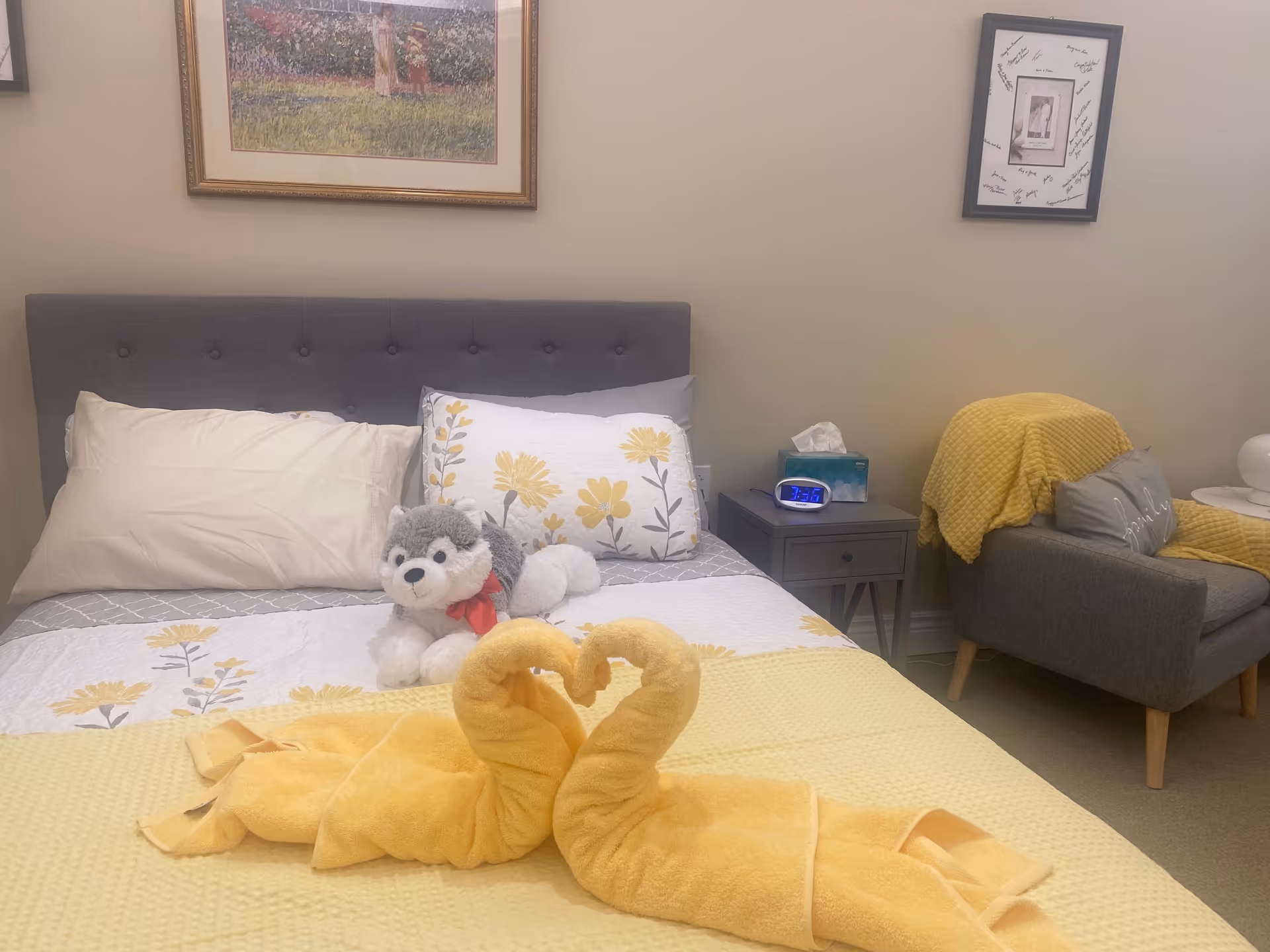 A cozy bedroom with a gray upholstered headboard, a bed with floral and plain pillows, a stuffed husky toy, and two yellow towel swans forming a heart shape on a yellow blanket. Next to the bed is a gray nightstand with a digital clock and a tissue box. A gray armchair with a yellow blanket and a pillow is in the corner. Two framed pictures hang on the beige walls.