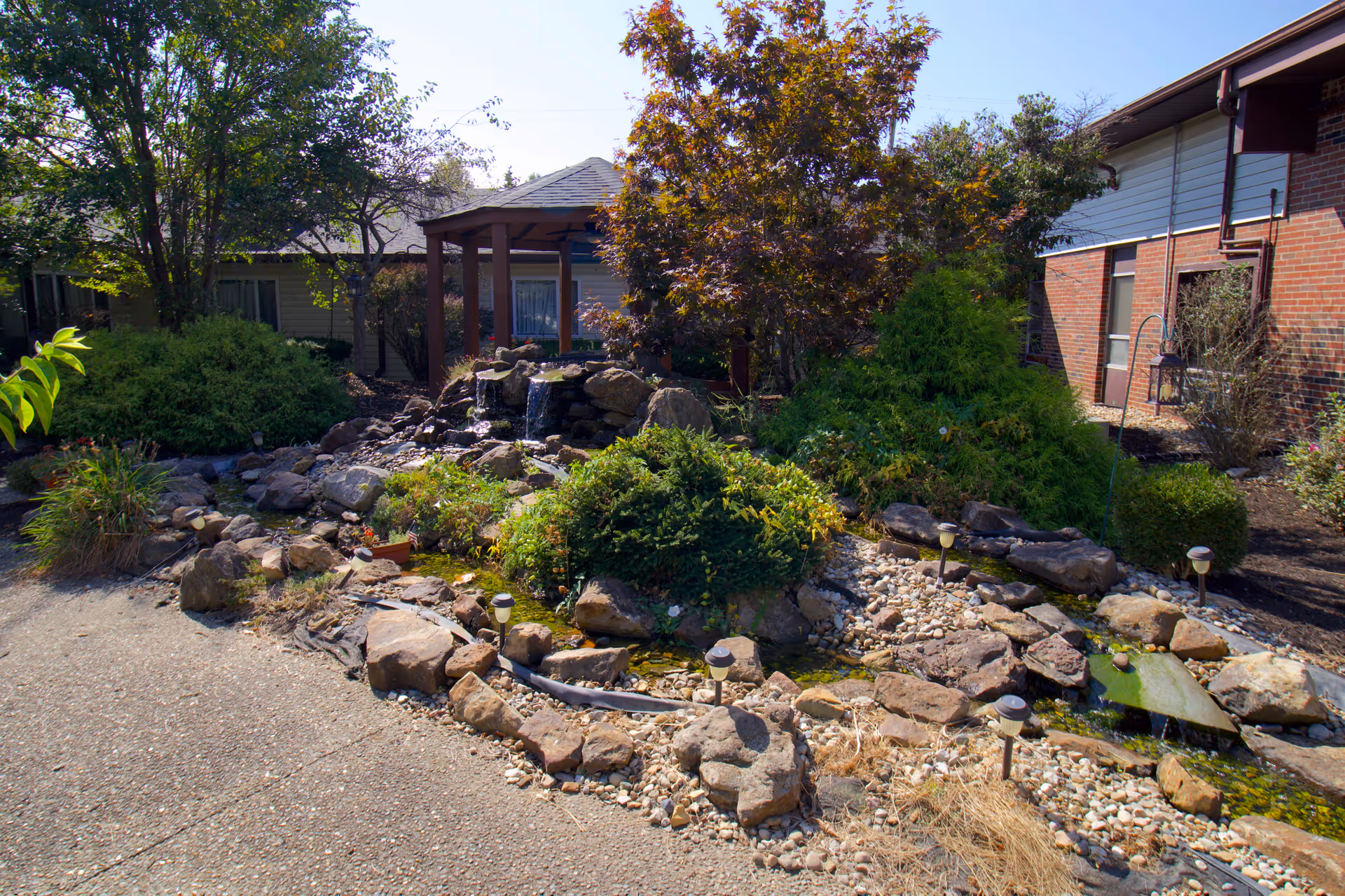 Outdoor garden area at Majestic Care of Bloomington featuring a small rock waterfall, various shrubs and trees, a paved walkway, and a wooden gazebo in the background next to brick and siding buildings.