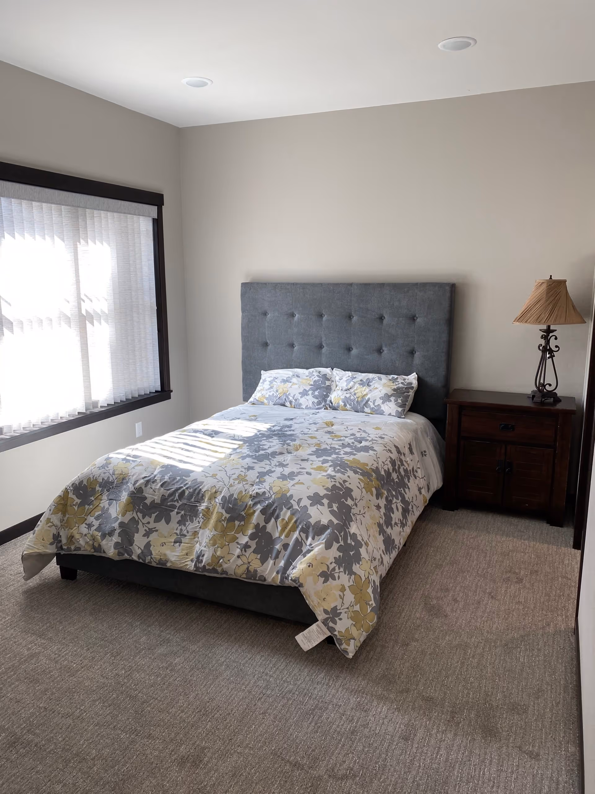 A bedroom with a bed featuring a gray tufted headboard and floral-patterned bedding in shades of gray and yellow. Next to the bed is a dark wooden nightstand with a decorative lamp. A large window with vertical blinds allows natural light to fill the room. The walls are painted light beige and the floor is carpeted in a neutral tone.