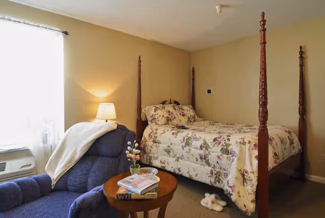 Cozy bedroom with a four-poster bed in floral bedding, a blue armchair, and a small round table with magazines.