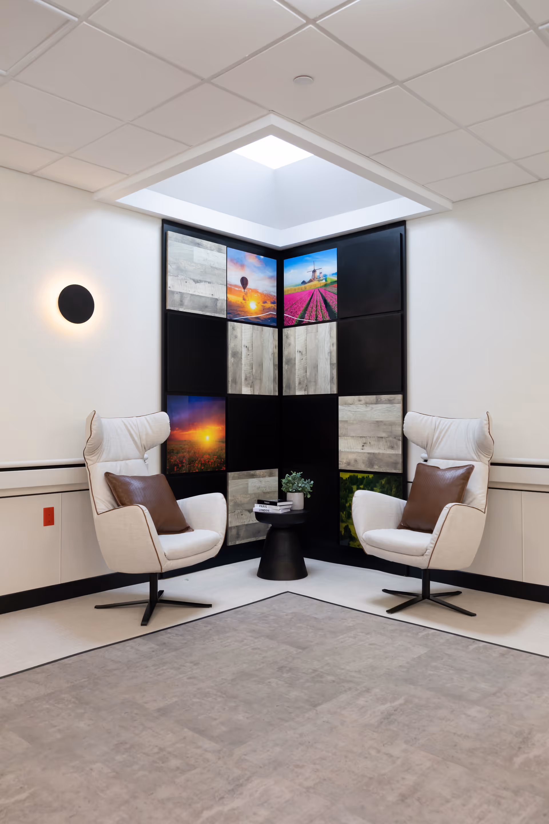 A cozy corner seating area with two modern white armchairs featuring brown cushions, positioned around a small black round table with a plant and books. The corner wall has a decorative panel with a mix of wood textures and colorful landscape images. The ceiling has a recessed skylight providing natural light, and a round wall light is mounted on the left wall.