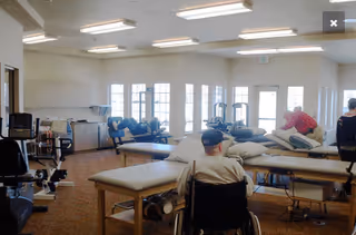 A rehabilitation room with several therapy tables and exercise equipment. There are three people visible, one sitting in a wheelchair facing away from the camera, and two others engaged in activities near the therapy tables. The room has large windows letting in natural light and bright ceiling lights.