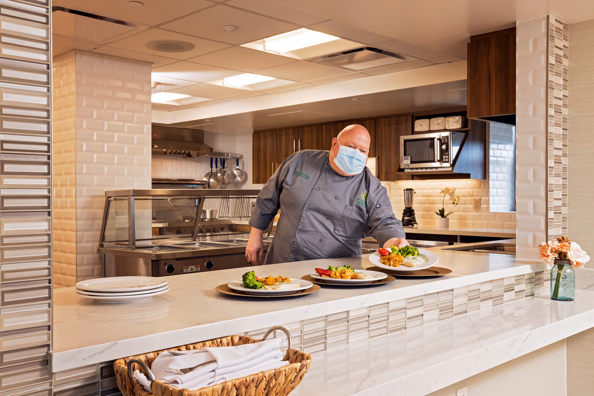 A chef wearing a face mask and gray uniform places plates of food on a white countertop in a modern kitchen with wooden cabinets and stainless steel appliances.