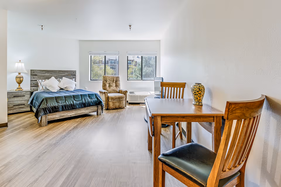 A bright senior living studio apartment with a bed, two pillows, a nightstand with a lamp, a recliner chair, two windows with a view of trees and buildings, a small wooden dining table with two chairs, and a decorative vase on the table.