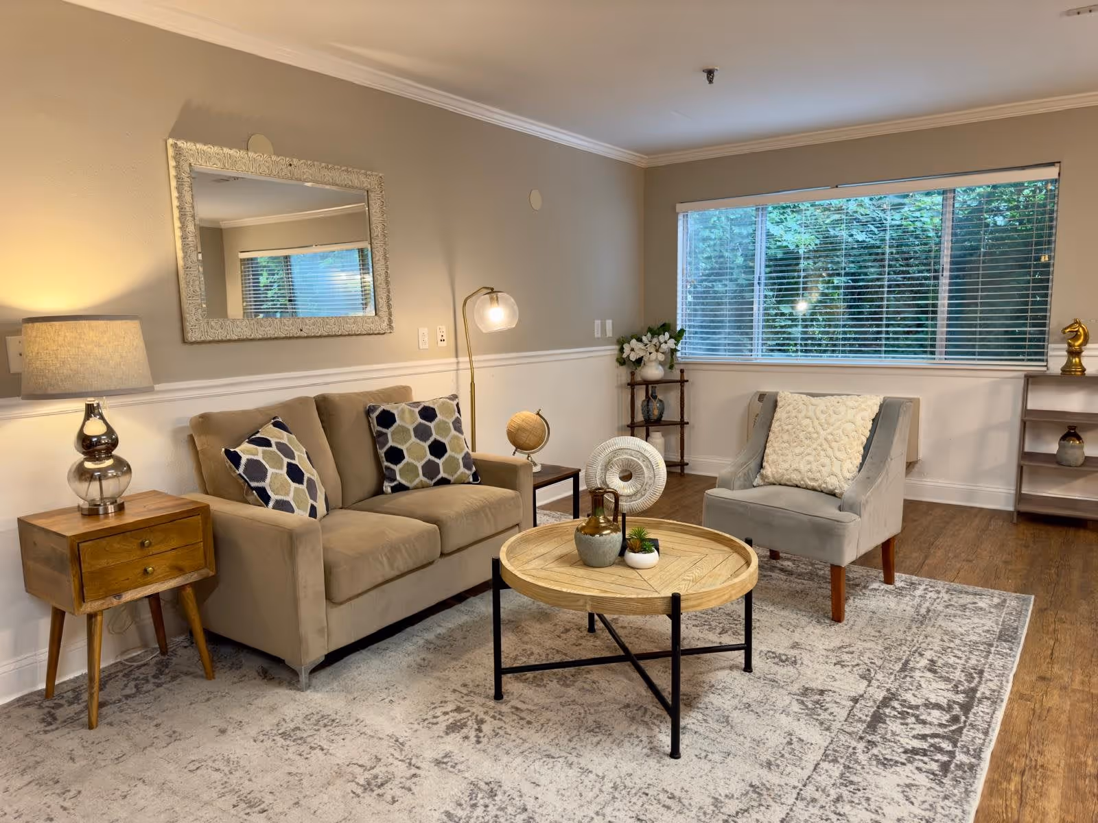 A cozy living room with a beige sofa adorned with two patterned pillows, a wooden side table with a lamp, a round wooden coffee table with decorative items, a gray armchair with a textured cushion, a large window with blinds showing greenery outside, and a small wooden shelf with decorative pieces.