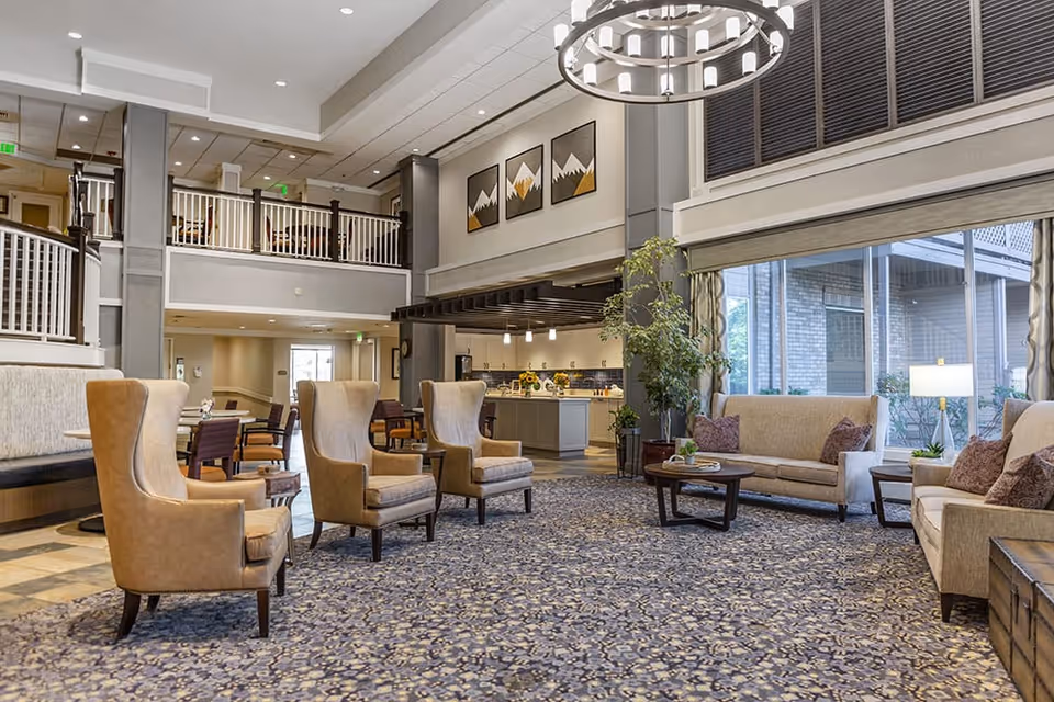 A spacious and well-lit living room area in a senior living facility with beige armchairs and sofas arranged around a patterned carpet. There is a large window on the right side letting in natural light, a modern chandelier hanging from the ceiling, and a kitchen area visible in the background. The room features neutral tones and decorative artwork on the walls.