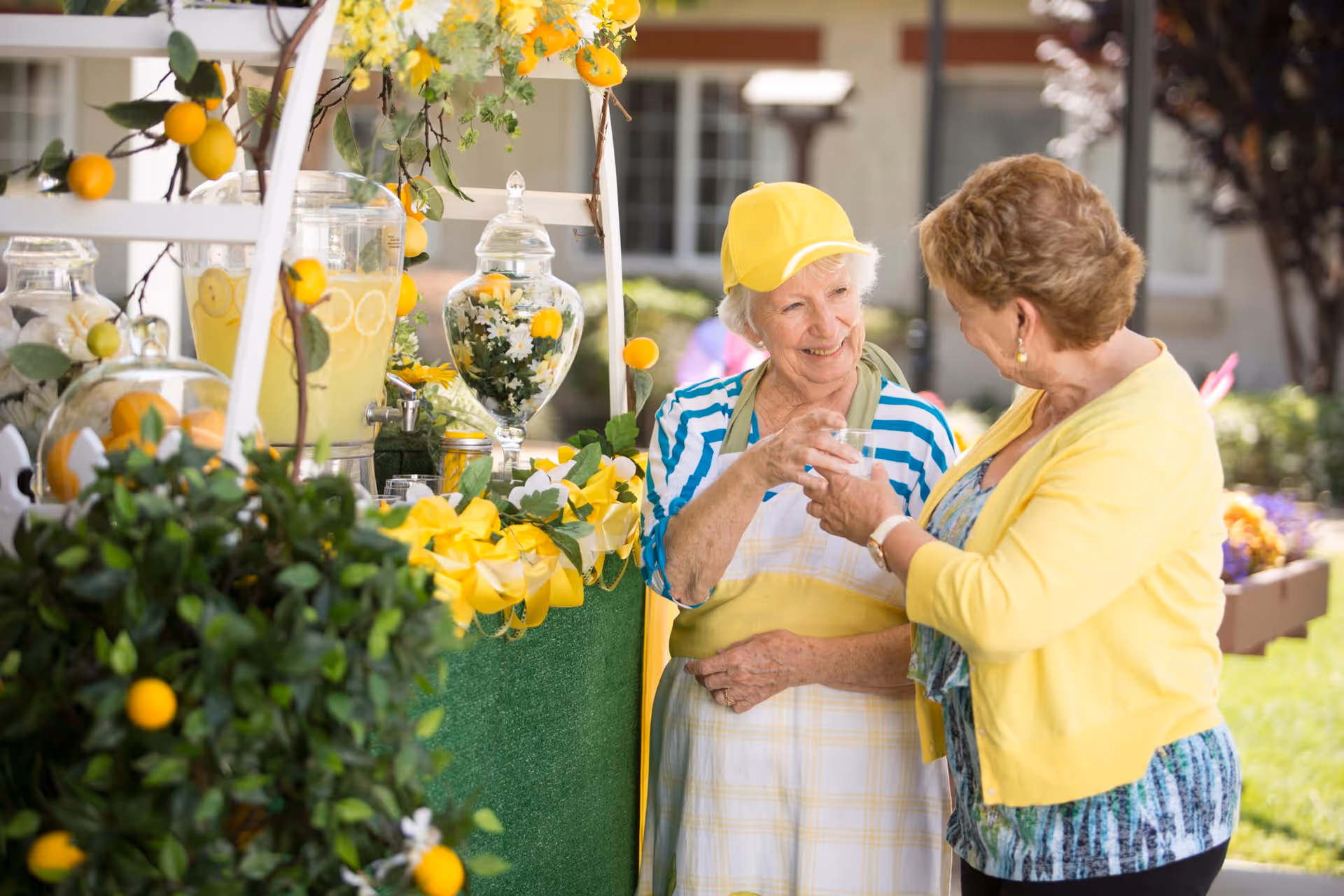 Two elderly women smiling and interacting at an outdoor lemonade stand decorated with yellow flowers and greenery. One woman is wearing a yellow cap and apron, holding a glass of lemonade, while the other woman, dressed in a yellow cardigan, is handing her the glass. The background shows a residential building and garden area.