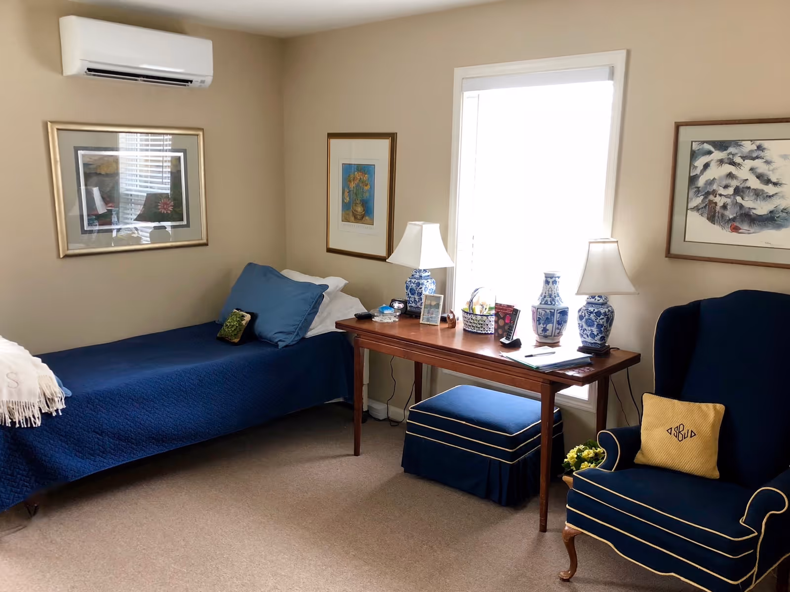 Neatly furnished single-occupancy bedroom with a blue-covered bed, wooden desk with lamps under a window, and a blue armchair.