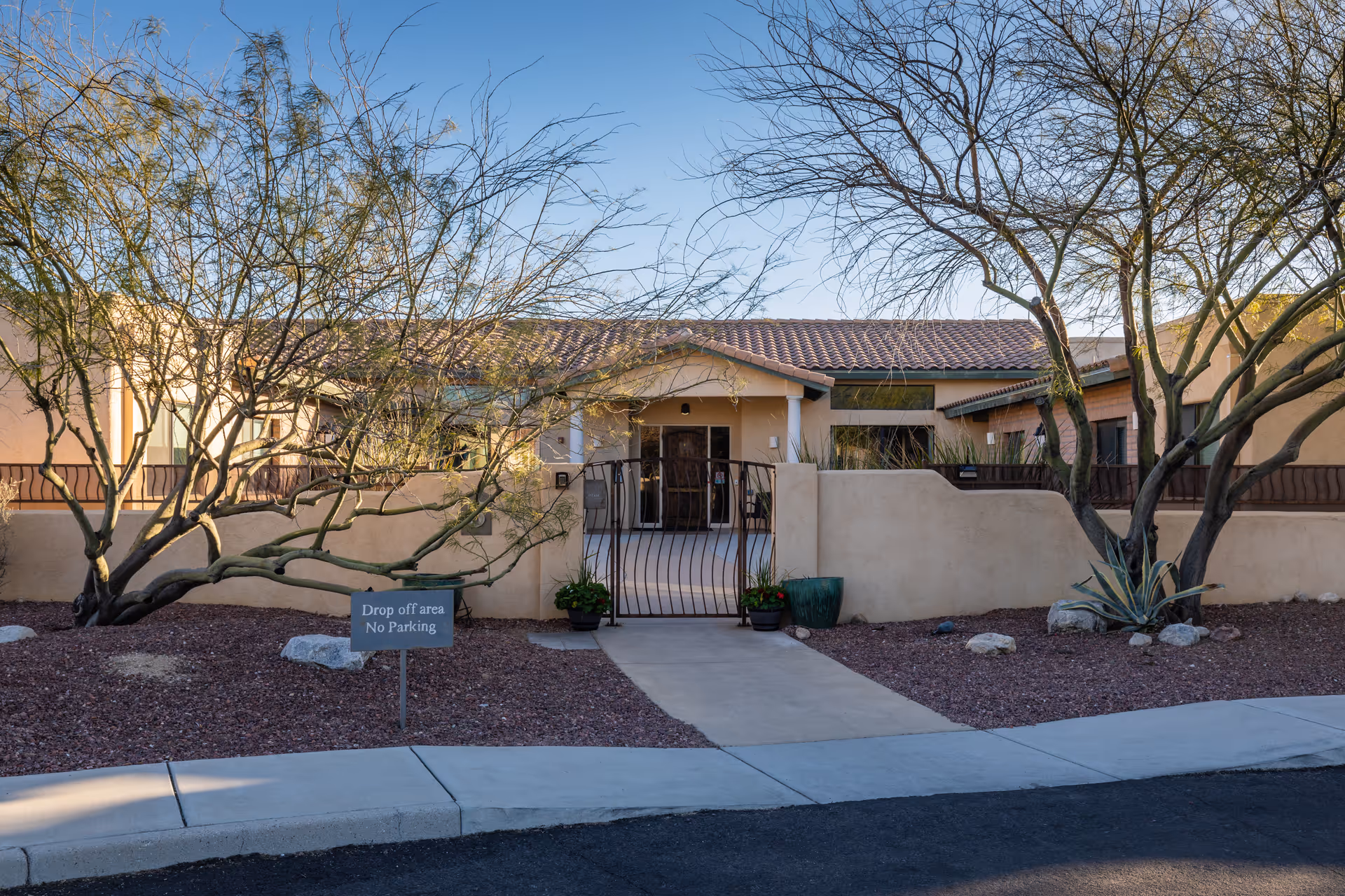 Exterior view of BridgeWater Assisted Living - Tucson showing a single-story building with a tiled roof, beige stucco walls, and a gated entrance. There are desert landscaping elements including leafless trees, rocks, and gravel. A sign near the entrance reads 'Drop off area No Parking'.