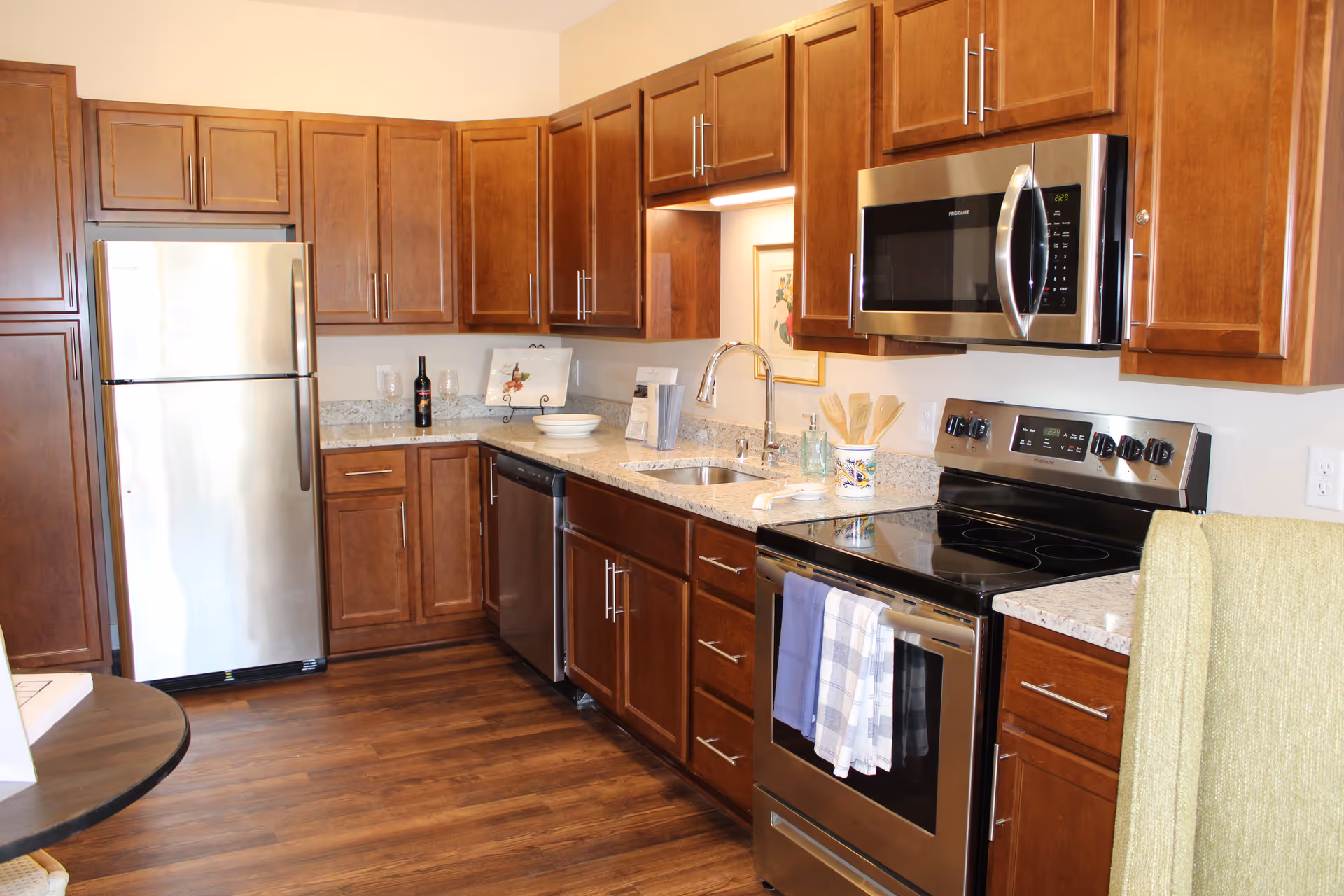 A modern kitchen with wooden cabinets, stainless steel refrigerator, dishwasher, microwave, and electric stove. The countertop is granite with a sink and various kitchen items including a bottle of wine, bowls, and utensils. The floor is wooden and there is a small round table partially visible on the left.