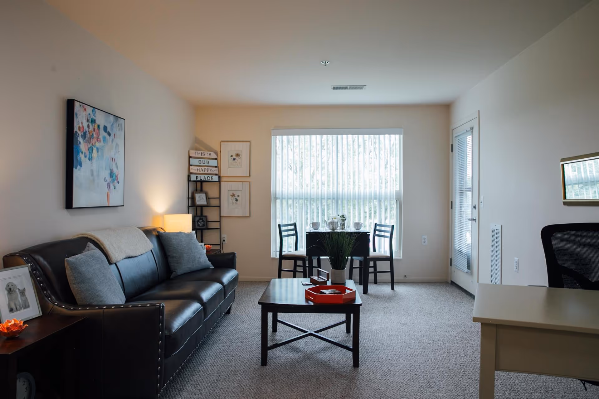 Bright furnished living room with a leather sofa, coffee table, small dining set by a large window, and a desk on the right.