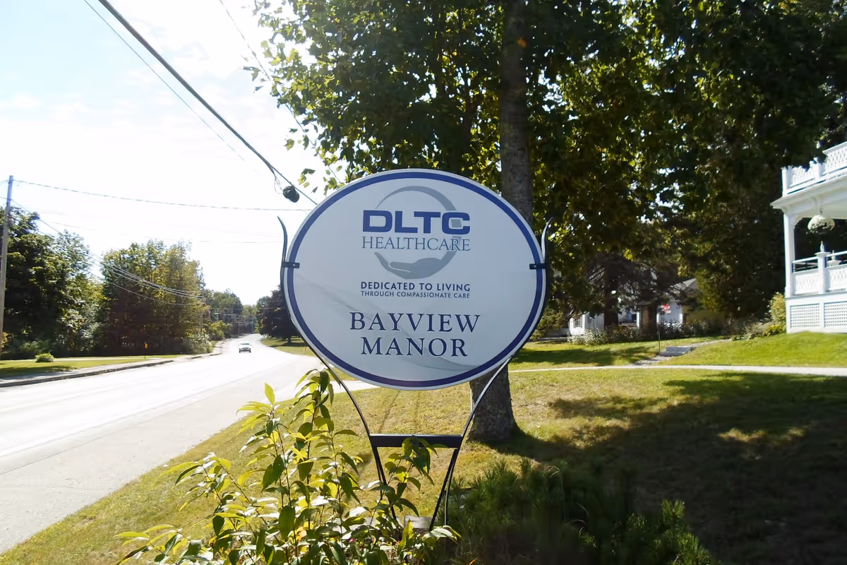 Outdoor view of a sign for Bayview Manor, a DLTC Healthcare facility, located on a grassy area next to a road with trees and houses in the background.