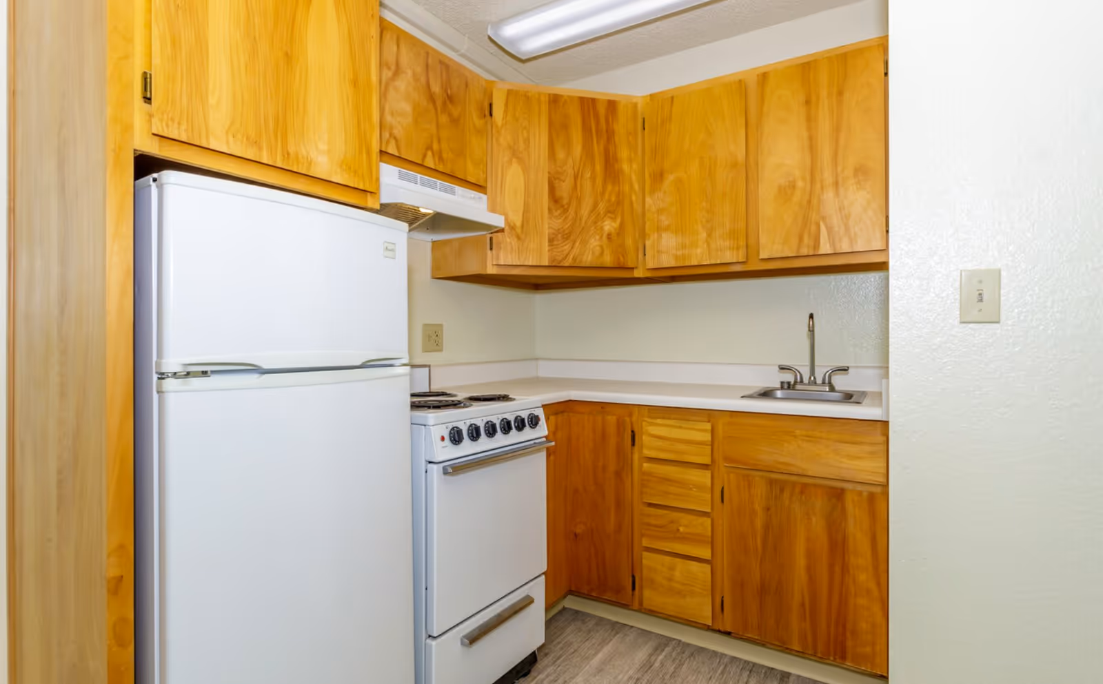 Small kitchen area with wooden cabinets, a white refrigerator, a white stove with oven, a stainless steel sink, and a countertop. The walls are light-colored and the floor has a wood-like finish.