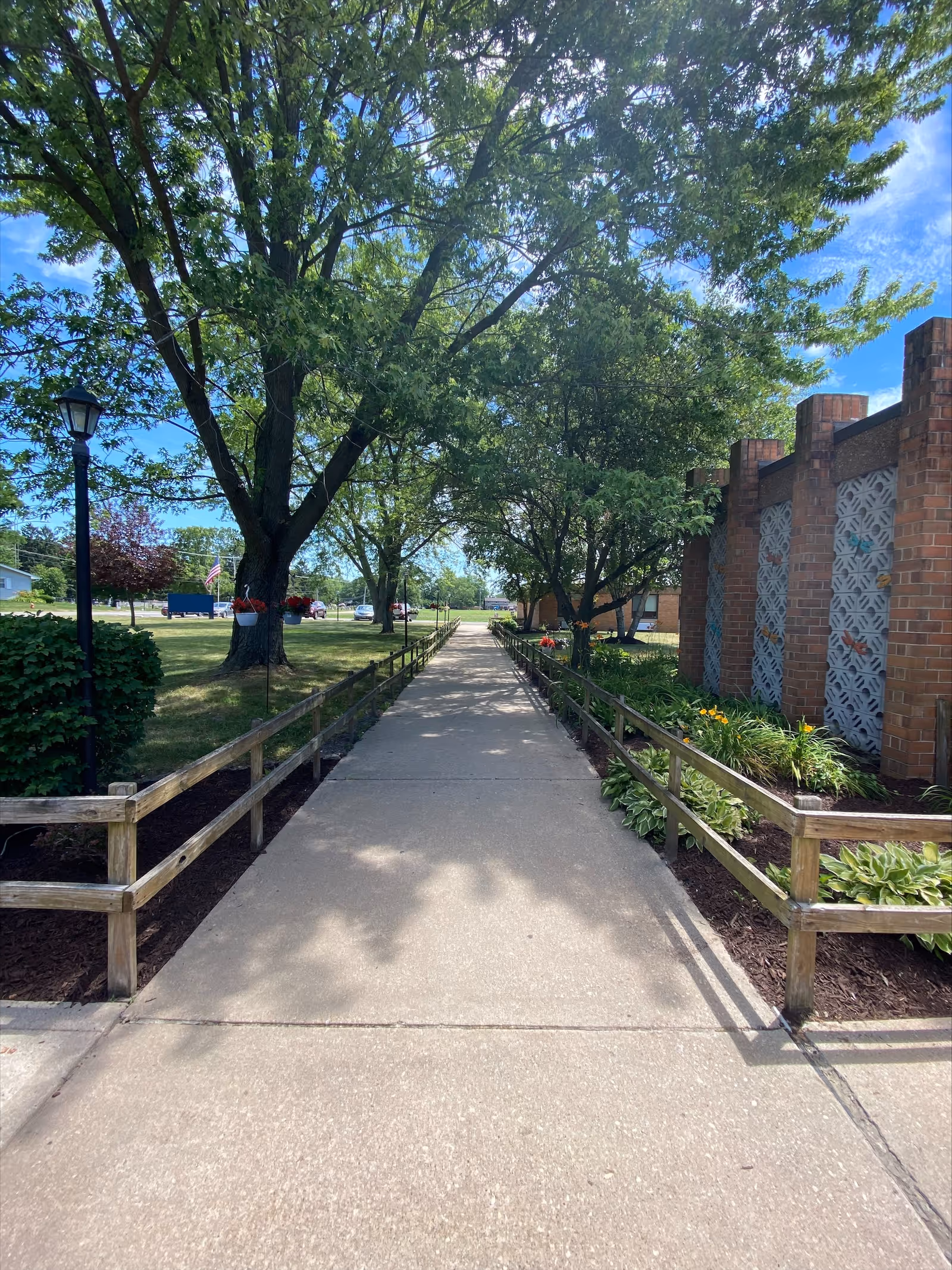 A concrete walkway lined with wooden railings on both sides, leading through a garden area with green trees and plants. On the right side, there is a brick building with decorative concrete blocks. The sky is blue with some clouds, and there is a lamp post on the left side of the walkway.