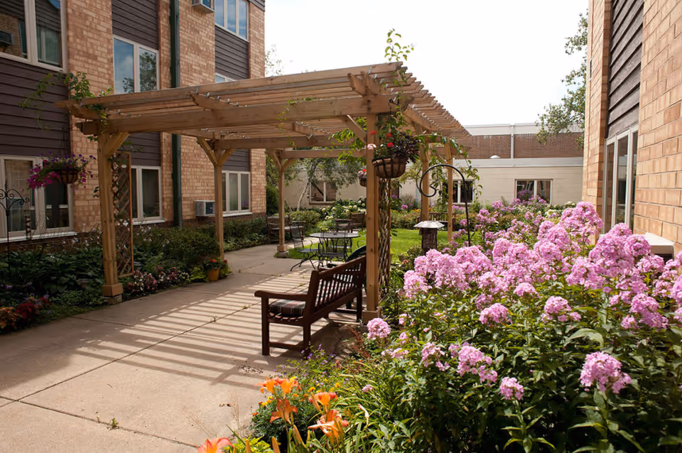 Sunny courtyard with a wooden pergola, bench and patio seating surrounded by blooming flower beds between brick buildings.